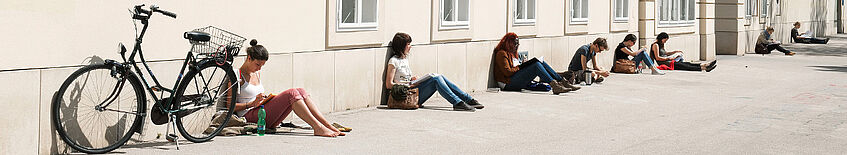 Abbildung: Campus der Uni Wien an einem sonnigen Sommertag, Studierende sitzen auf dem Boden angelehnt an eine Hauswand.